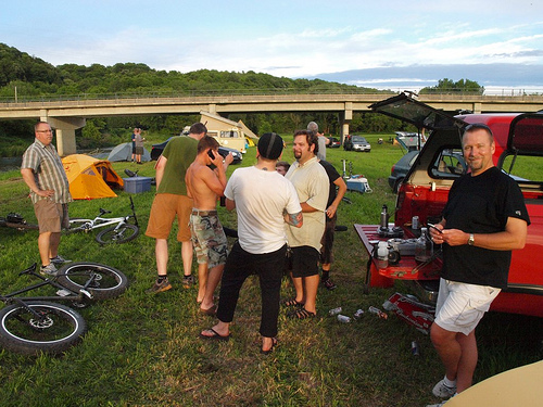 People standing around on a grass field with campsites, bikes and vehicles, with a bridge and trees in the background